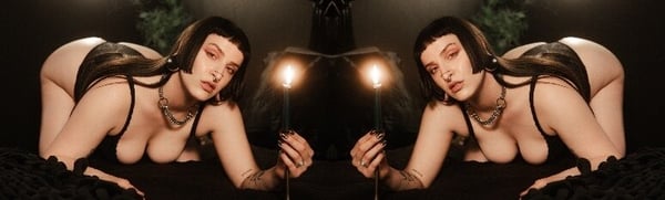 A woman in dark lingerie poses symmetrically on a bed with candles and plants in a dimly lit gothic setting.