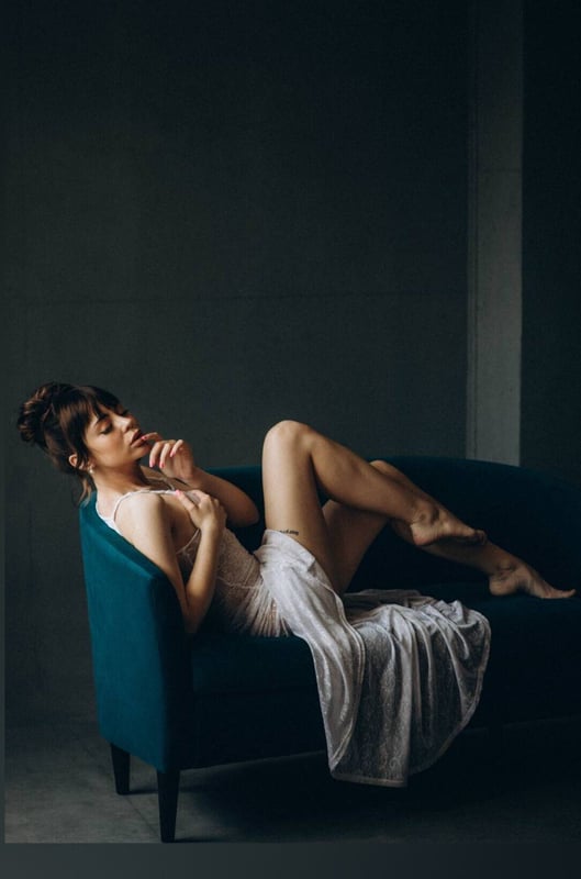 A woman reclining on a teal sofa in a sheer white dress, posing thoughtfully with her hand near her face against a dark background.