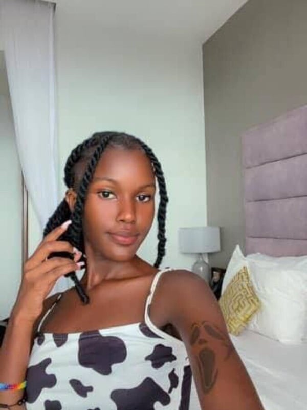 A woman with braided dark hair takes a selfie in a black and white cow print top while sitting on a bed in a bedroom.