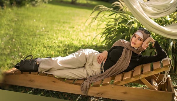 A woman in modest attire with a headscarf reclines on a wooden lounge chair in a lush green garden setting with flowing white fabric nearby.