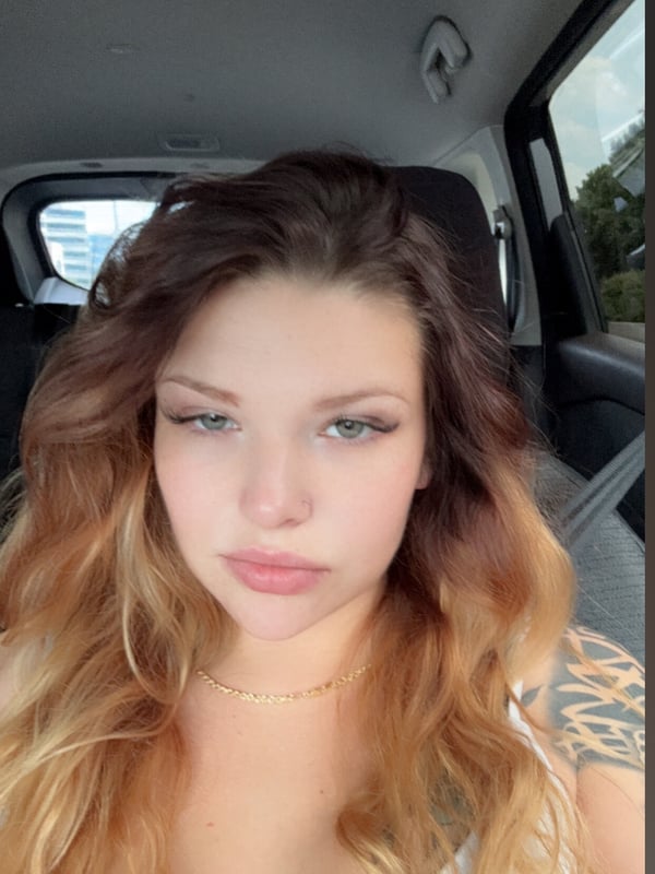 A young woman with wavy blonde hair and tattoos sitting in a vehicle, wearing a white top and gold necklace.