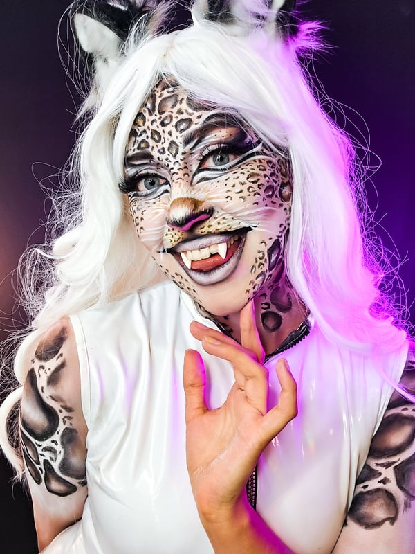 A person with intricate leopard face paint, white hair, cat ears, and glowing purple makeup, posing playfully against a dark background.