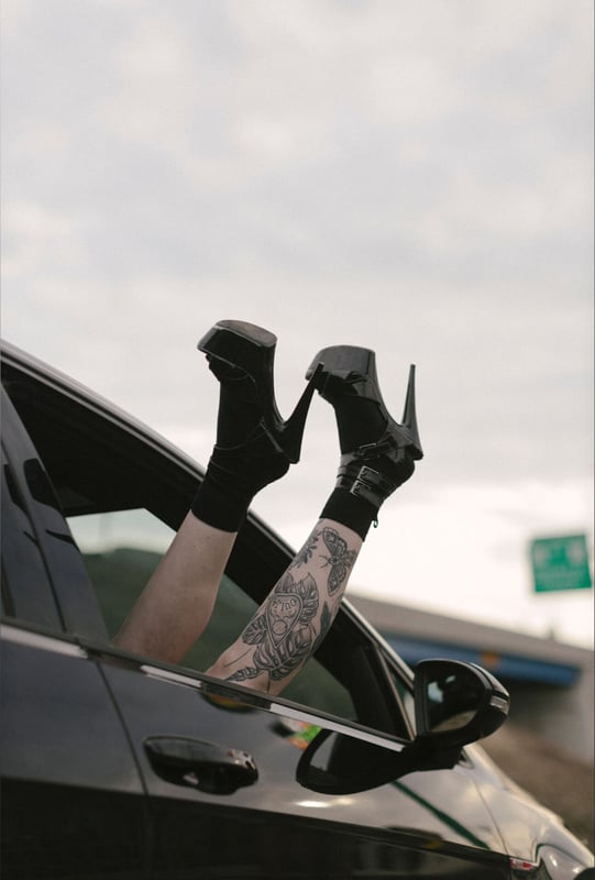 Person's legs in black high heels extending from a car window, with visible tattoos on the legs, against a cloudy sky and road sign.