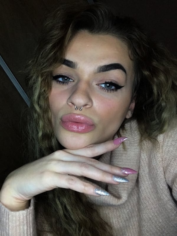 Young woman with wavy brunette hair and bold makeup, posing with her hand on her chin in a close-up selfie indoors.