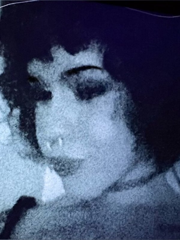Close-up portrait of a young woman with curly hair, piercings on her face, and a subtle expression in a dimly lit setting.