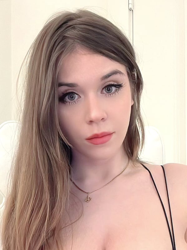 Young woman with long brown hair, posing with a subtle expression in a low-cut black top against a light indoor background.