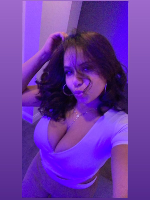 Young woman with wavy brown hair and hoop earrings posing confidently in a low-cut white top under purple lighting.