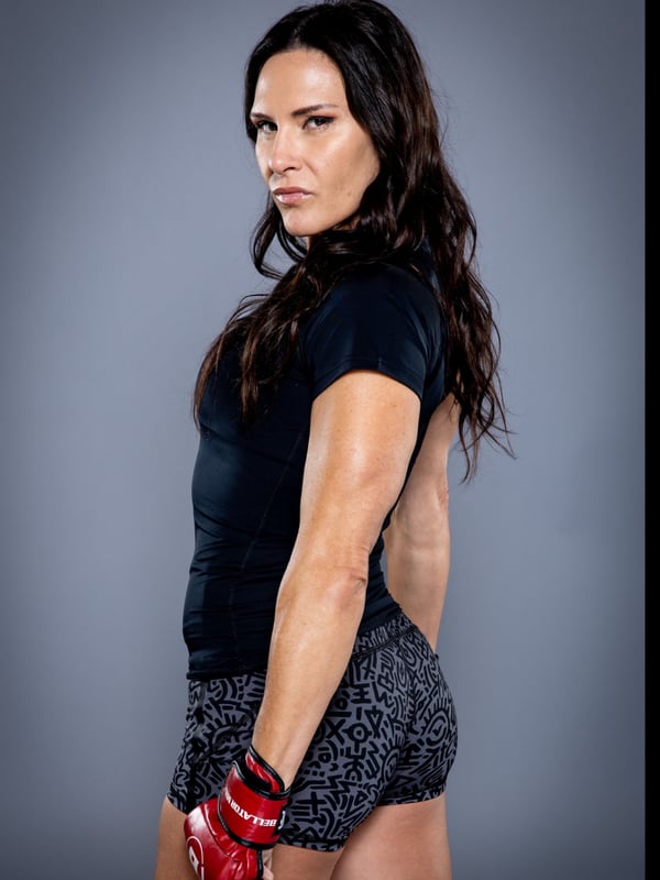 A muscular woman in a black tank top and shorts with red boxing gloves poses confidently against a gray background.