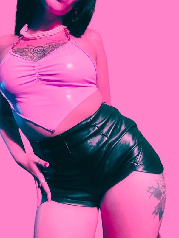 Woman posing confidently in shiny pink top and black shorts against a vibrant pink background.