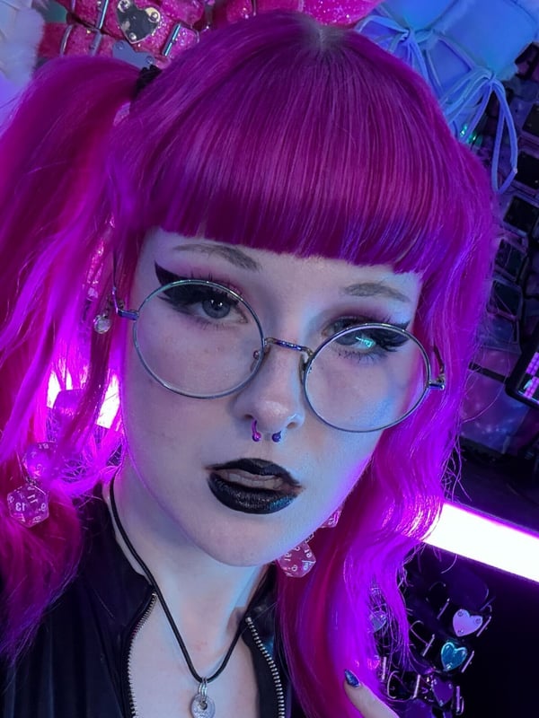A woman with pink hair in pigtails, glasses, and goth makeup posing in a vibrant neon-lit setting with decorative elements.