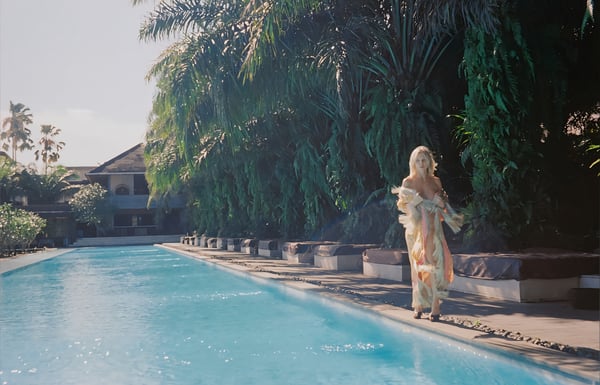 A blonde woman in a flowing dress walks by a tropical poolside with palm trees and a luxurious villa in the background.