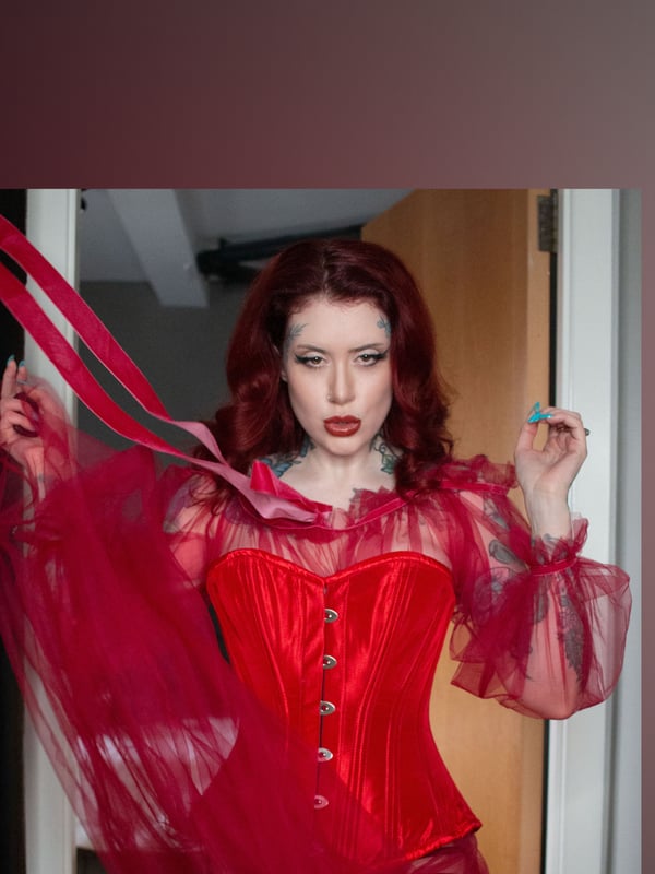 Woman with red hair holding a red ribbon, wearing a sheer red corset top near a door.