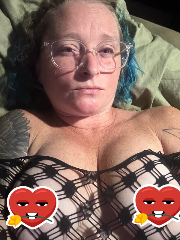 A woman with blue hair and glasses lies on a bed wearing a black lace top with heart emojis, smiling playfully.