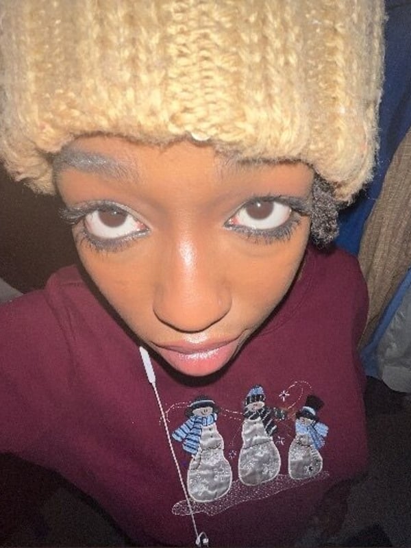 Young woman with blonde hair wearing a knitted beanie and a maroon sweatshirt with snowman designs, posing indoors with clothes hanging in the background.