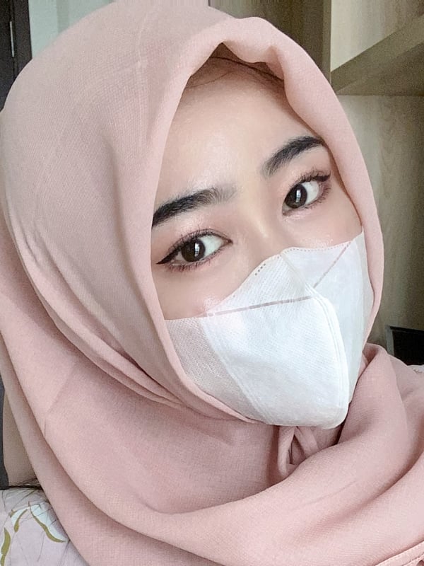 A close-up of a woman wearing a light pink headscarf and white face mask, looking toward the camera.