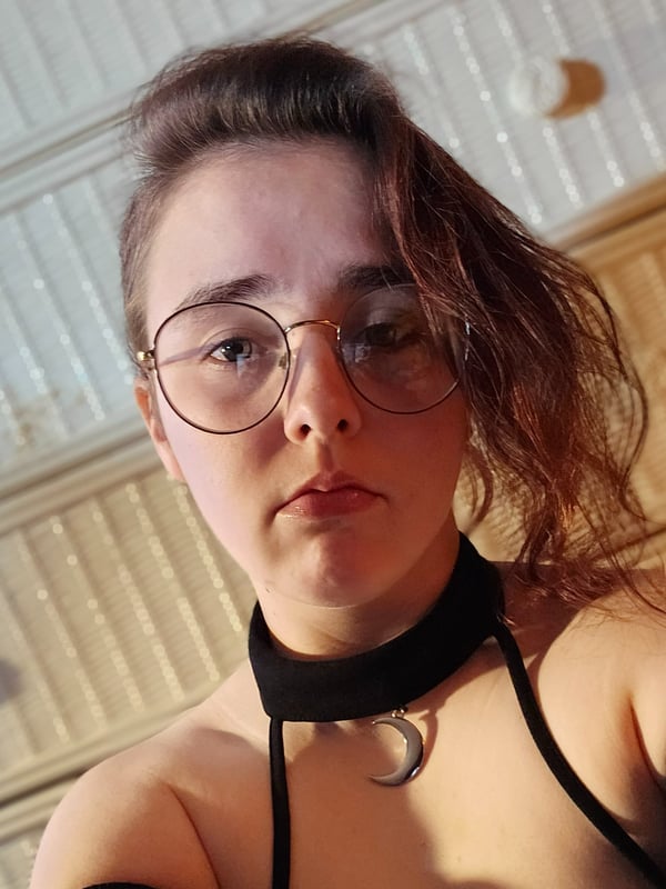 Young woman with glasses and wavy brown hair, wearing a black choker and arm sleeves, posing in front of wooden drawers.