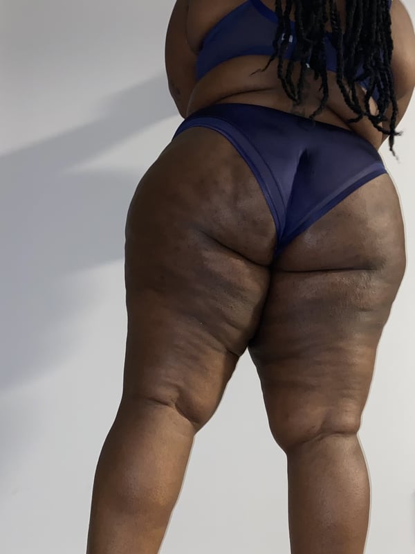 A woman standing viewed from behind, wearing blue lingerie, with braided hair and curvy figure.