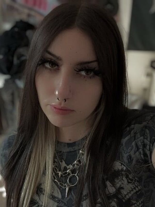 A young woman with long dark hair and piercings taking a selfie in a bedroom, wearing a patterned top with tattoos visible on her arm.