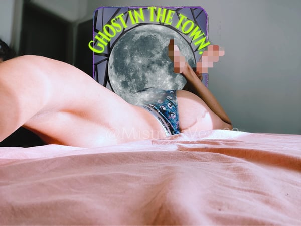 A person reclining on a bed with legs extended, positioned beside a vibrant graphic poster featuring a moon and the text 'Ghost in the Town' against a dimly lit room.