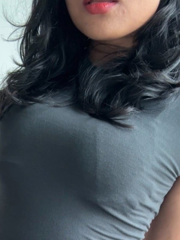 Young woman with long wavy dark hair and red lips, wearing a gray top, smiling softly against a neutral background.