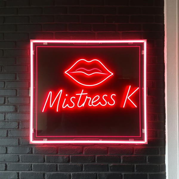Neon sign featuring red lips and the text 'Mistress K' framed against a dark brick wall.
