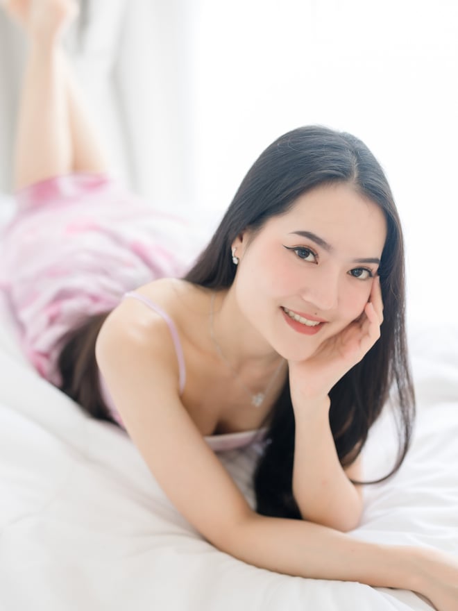 A smiling woman with long dark hair posing on a bed in a softly lit room.