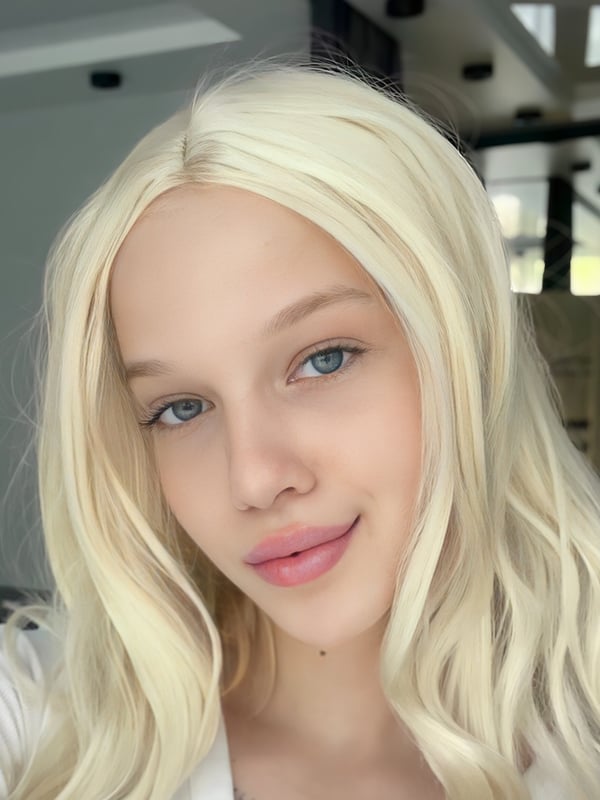 Close-up portrait of a young woman with long blonde hair and blue eyes smiling softly while wearing a white top in an indoor setting.