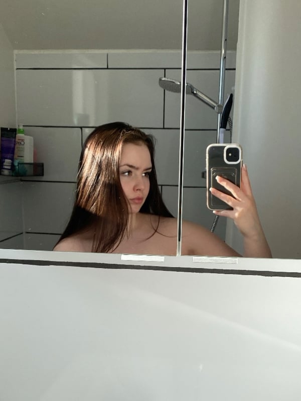 A young woman taking a selfie in a bathroom with white tiled walls and a glass shower door, holding a smartphone.