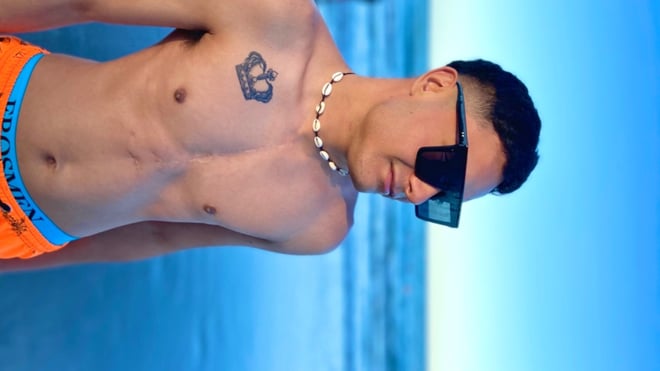 A young man with sunglasses and a necklace poses shirtless in orange swim trunks on a beach, showing a crown tattoo on his chest.