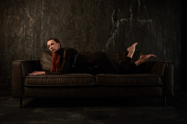 A woman with red hair lounging on a dark leather couch against a textured gray wall, wearing black clothing and a red scarf.