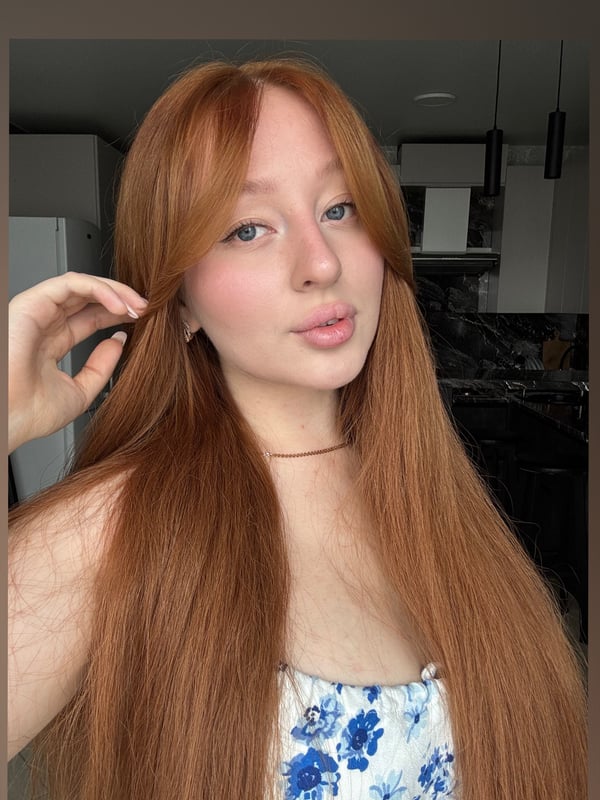 A young woman with long red hair and freckles smiling playfully while posing in a kitchen setting.