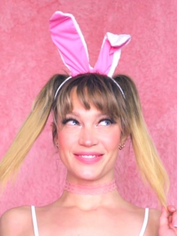 A woman with blonde pigtails and pink bunny ears smiling at the camera, wearing a choker against a pink background.
