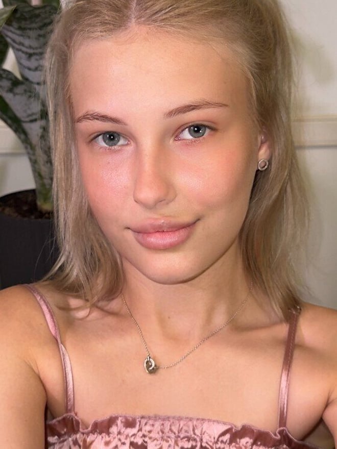 A close-up portrait of a young woman with blonde hair in a ponytail, wearing a pink satin top and a delicate necklace.