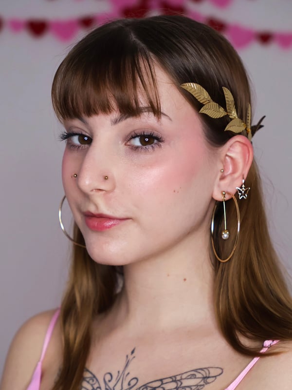 A person with long brown hair adorned with gold leaf accessories, smiling softly against a pink garland background.