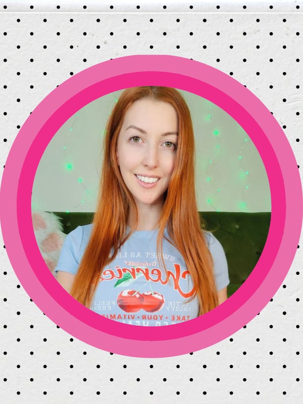 Close-up of a smiling redheaded woman with long hair, wearing a casual t-shirt, framed in a pink circle with sparkles.
