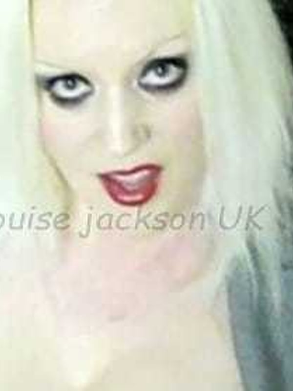 Close-up portrait of a woman with long blonde hair, dramatic makeup, and red lipstick, smiling playfully against a dark background.