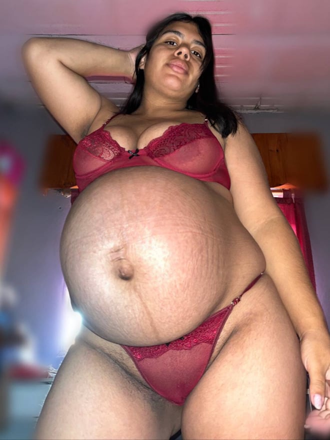 A pregnant woman standing indoors, wearing a red lace lingerie set and posing confidently.
