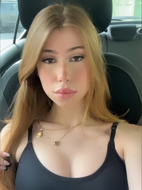 A young woman with long blonde hair sits in a car seat, wearing a black top, with a subtle smile and natural makeup.