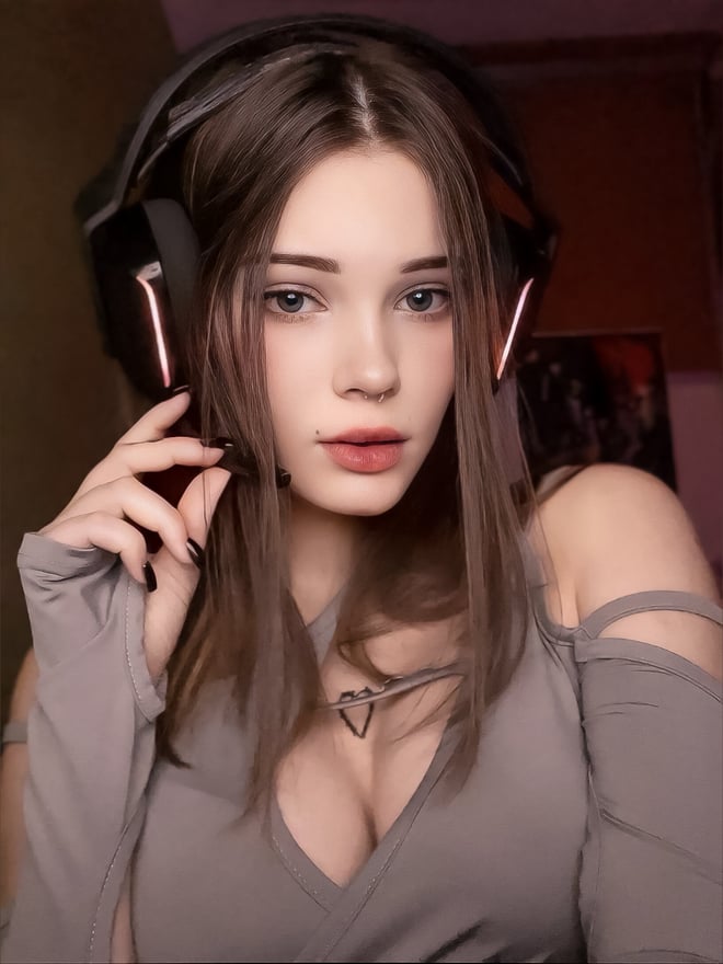 A young woman with long brown hair wearing headphones and a grey top, posing indoors.