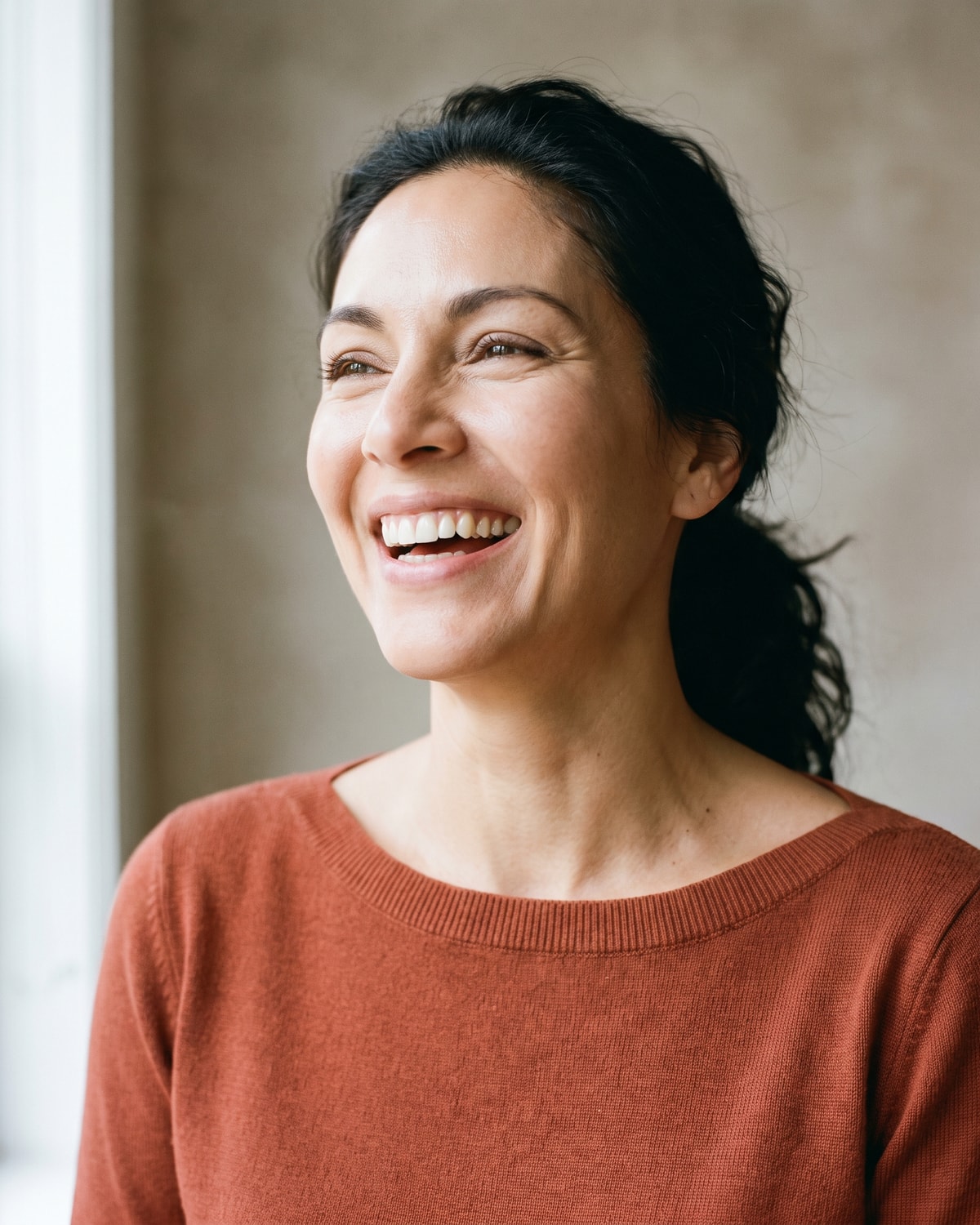 Woman in her forties laughing openly with her head tipped back, lit by broad soft daylight against a pale greige wall.
