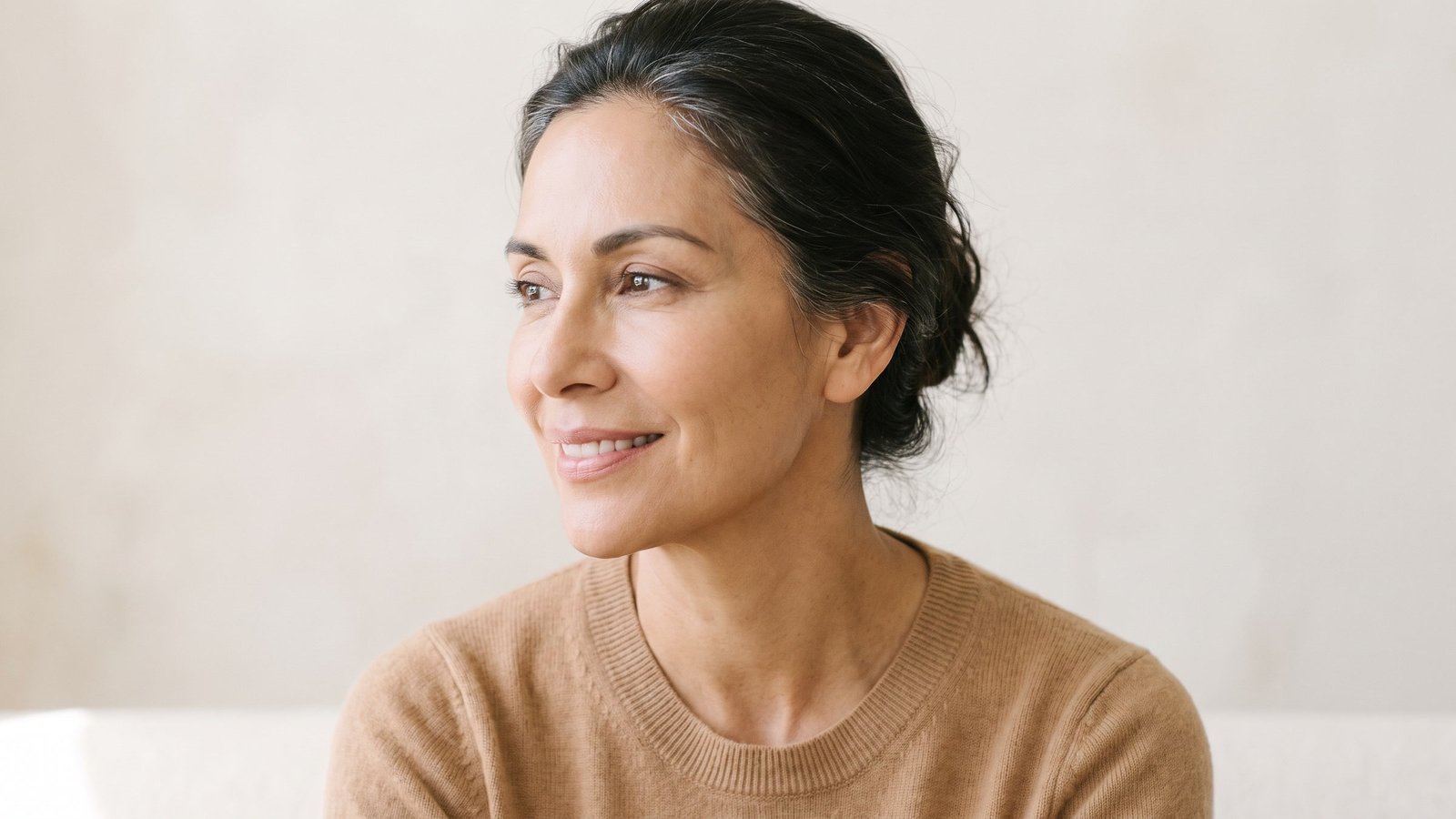 Latina woman in her fifties in three-quarter view with a warm slight smile, looking off to one side in soft open shade
