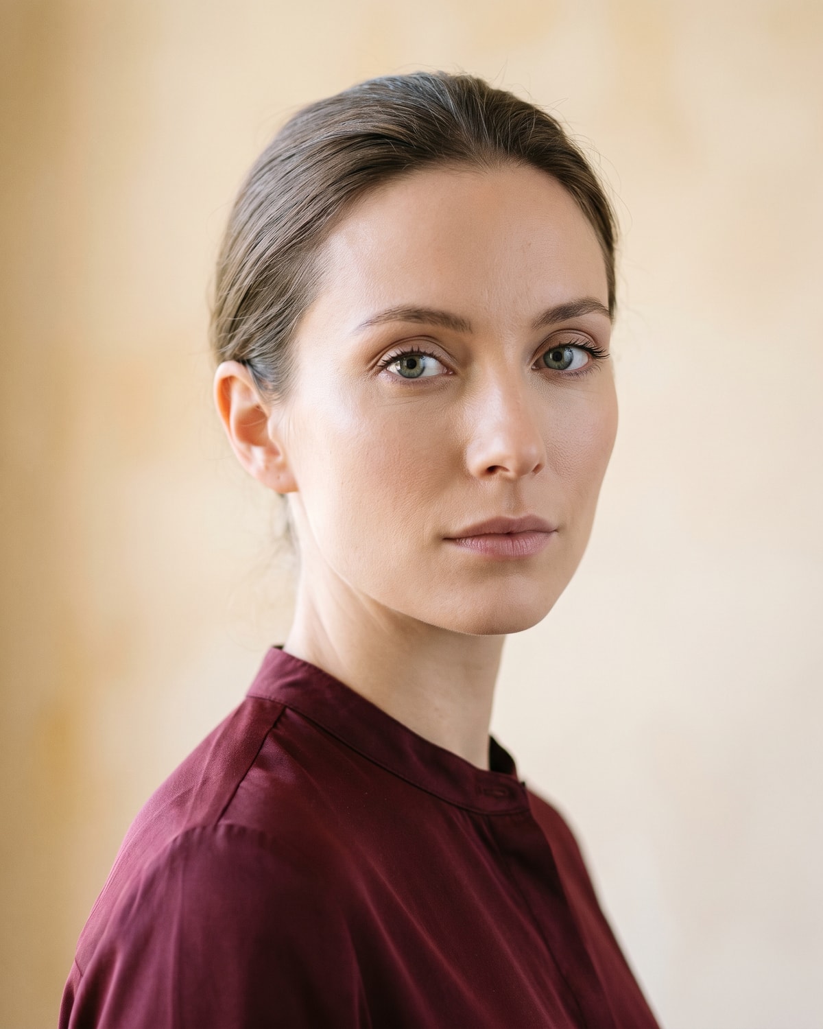 Woman in three-quarter view looking directly at the camera, eyes open and clear, in broad soft frontal light against a warm ochre-cream wall.