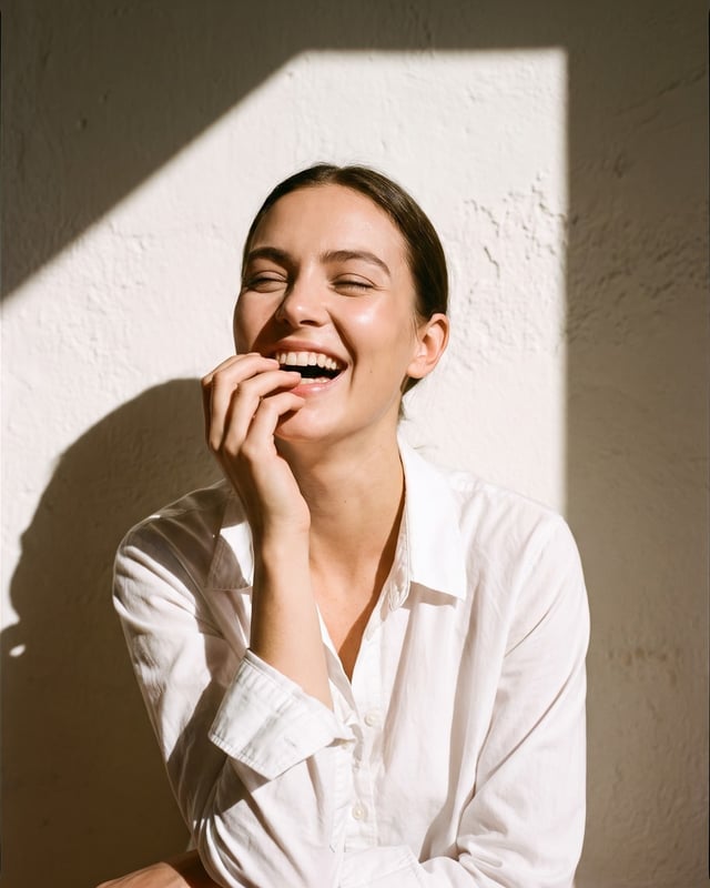 Woman laughing openly with her eyes crinkled shut, lit by bright bounced sunlight against a bone-white wall