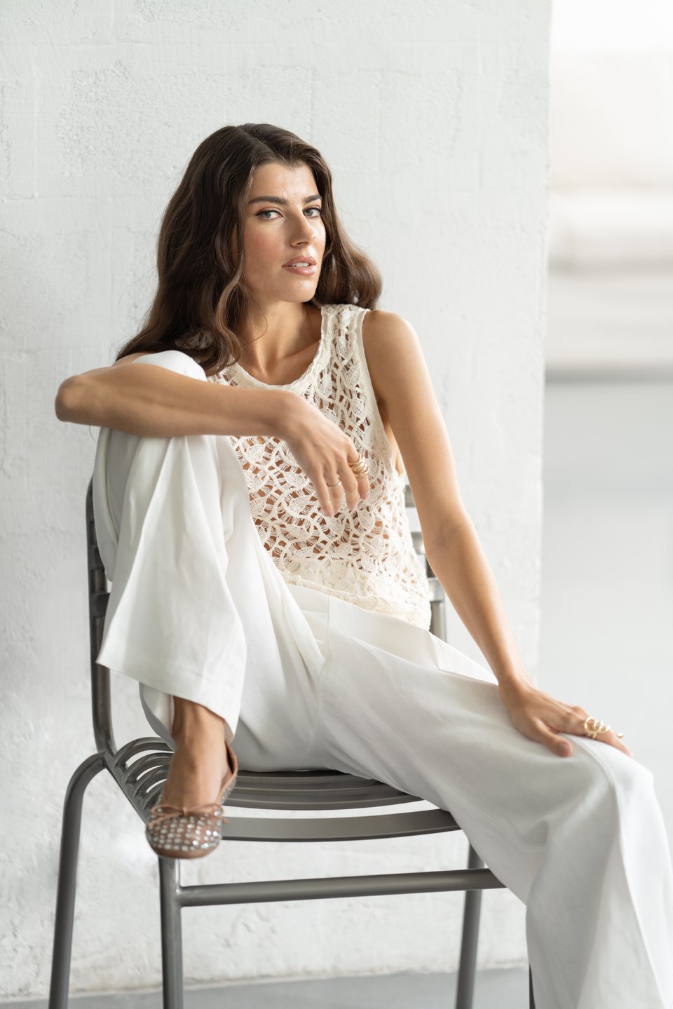 Editorial portrait of a woman in a white crochet top and linen pants seated on a metal chair in soft natural daylight