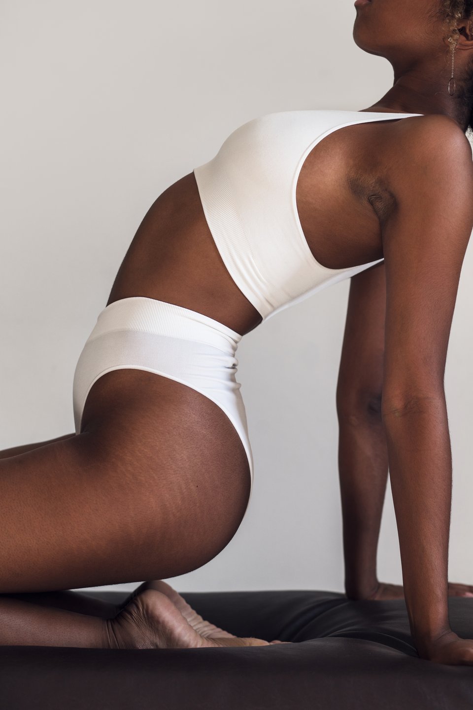 Editorial body silhouette of a woman in a cream sports bra and briefs in a low athletic crouch against a soft neutral backdrop