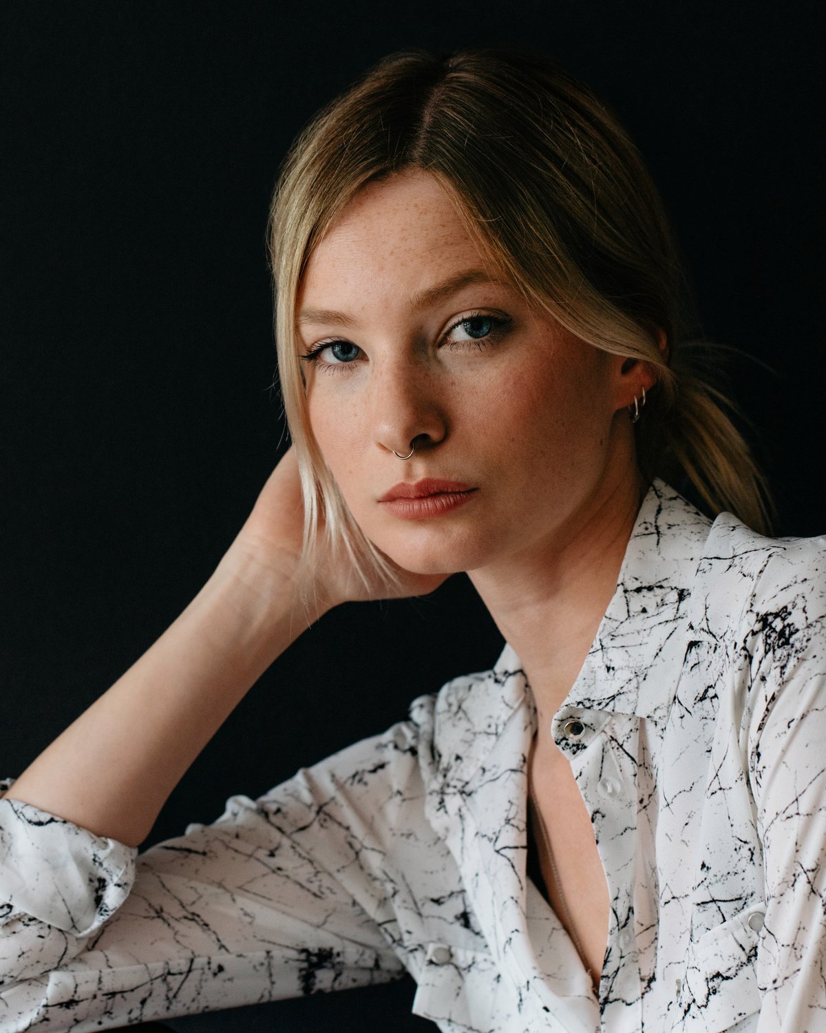 Editorial portrait of a young woman in a patterned blouse, hand at temple, against a dark backdrop