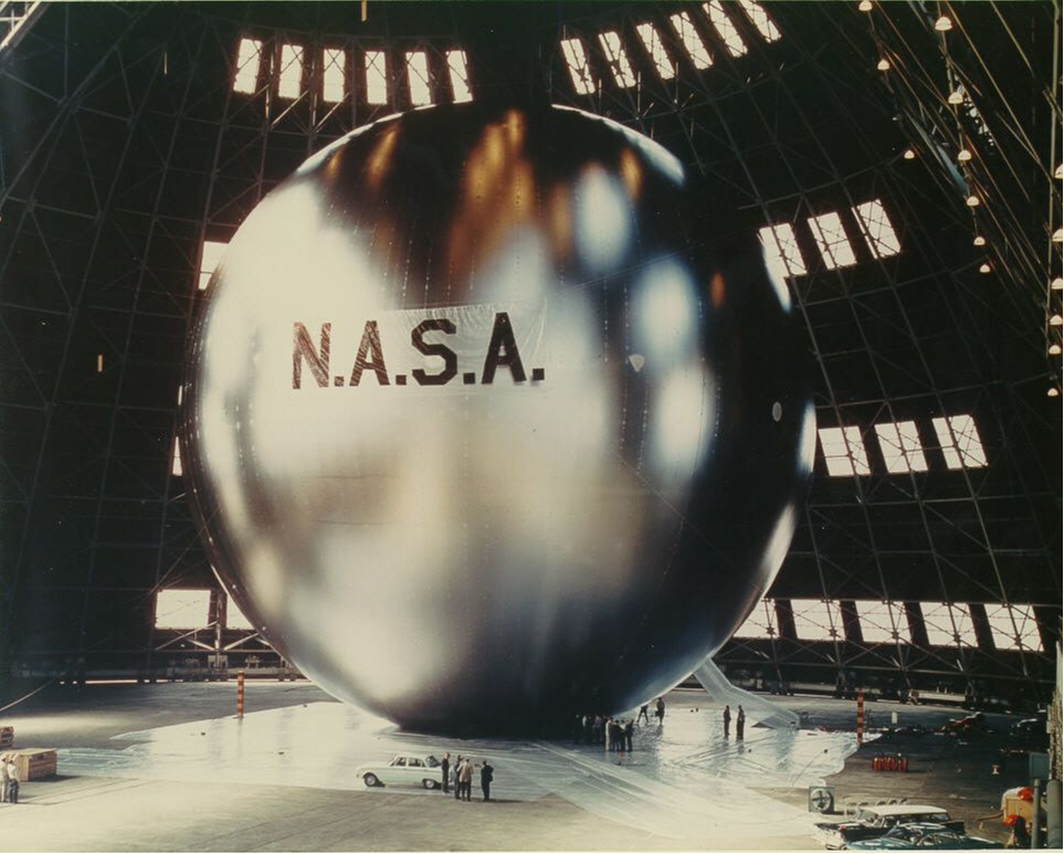 William J. O'Sullivan (sağda, ayakta) ve bir mühendis, sıkıca katlanmış ve paketlenmiş 12 feet çapında Beacon uyduyu içeren kapsülü inceliyor.