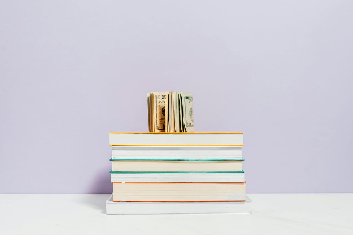 Stack of books with a stack of US dollar bills on top against a pastel background.