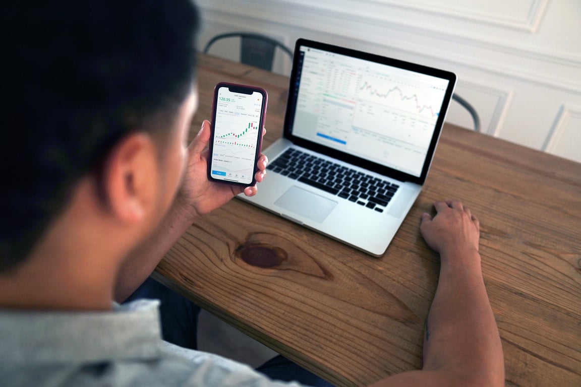 Individual reviewing stock market trends using a laptop and smartphone at a wooden table.