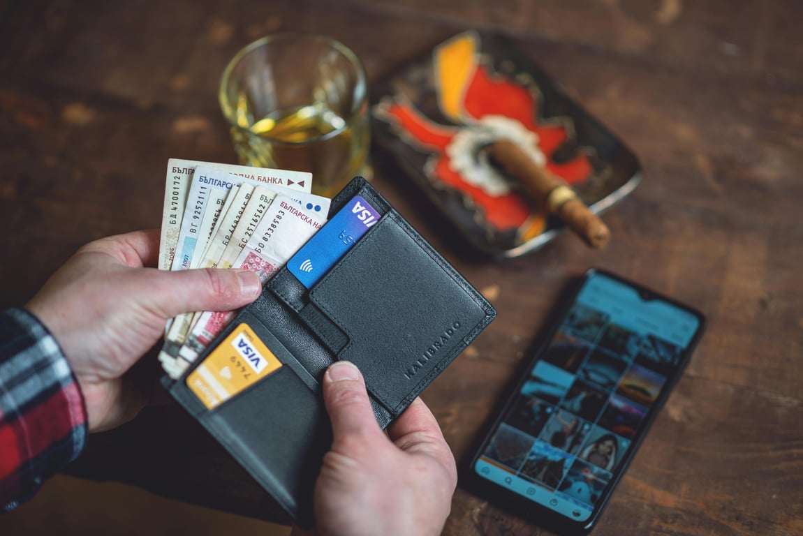 Close-up of hands holding a wallet with Bulgarian lev, credit cards, a smartphone, and a drink on a wooden table.